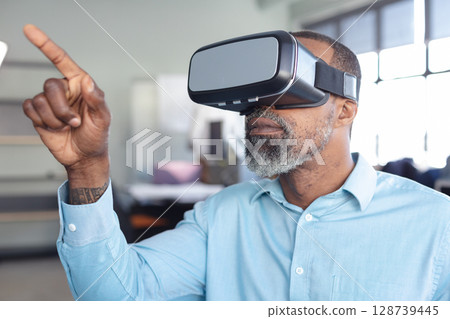 Standing African American man pointing forward in open-plan office, with virtual reality headset 128739445