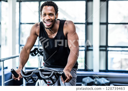 Pedaling African American man working out on stationary bike in modern gym, with dumbbells 128739463