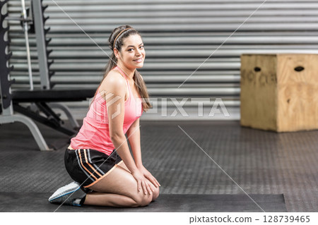 Kneeling female enthusiast exercising on dark gray mat in gym, with bench press and plyometric box 128739465