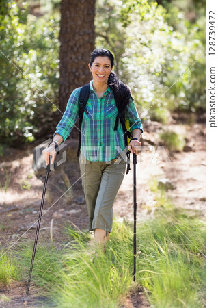 Hiking woman using trekking poles on forest trail, carrying backpack and wearing fitness tracker 128739472