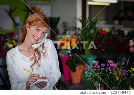 Red-haired woman florist writing on notepad while talking on phone at shop counter, copy space 128739475