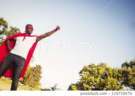 African American man standing heroically in park field, with red cape and wristbands, copy space 128739479