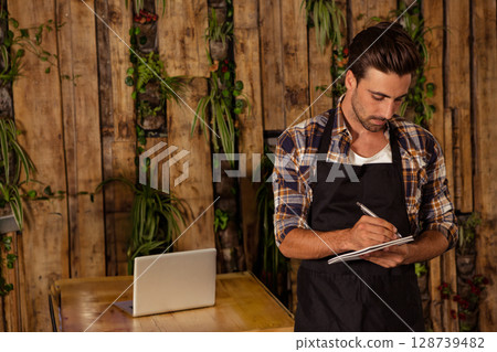 Male barista writing notes on notepad at wooden table in cafe, with laptop, plants, copy space 128739482
