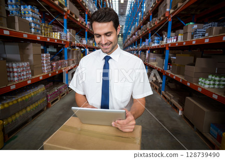 Scanning male warehouse worker reviewing inventory on tablet in warehouse aisle, with cardboard box 128739490