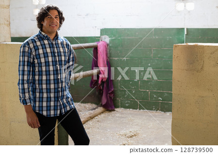 Standing man examining metal railings inside stable, with purple jacket, wood shavings, copy space 128739500