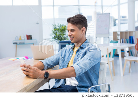 Smiling man holding smartphone and typing on laptop at office desk, with colorful sticky notes 128739503