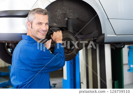 Mid adult male mechanic repairing brake assembly and smiling in garage, with silver car on lift Mid adult male mechanic repairing brake assembly and smiling in garage, with silver car on lift 128739510