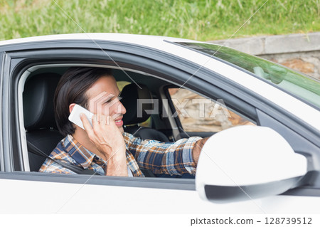 Talking man holding smartphone sitting in car beside stone wall, with seat belt, copy space Talking man holding smartphone sitting in car beside stone wall, with seat belt, copy space 128739512