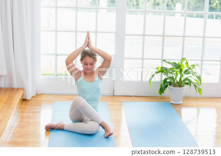 Stretching mature woman sitting cross-legged on blue yoga mats in bright studio, with green plant Stretching mature woman sitting cross-legged on blue yoga mats in bright studio, with green plant 128739513
