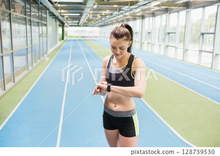 Standing woman checking smartwatch on blue and green track beside glass wall under metal beams 128739530