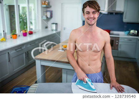 Shirtless man ironing shirt on ironing board at home kitchen island, with steam iron, copy space 128739540