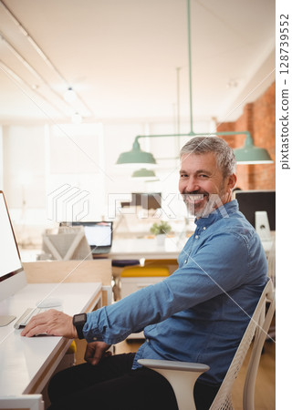 Sitting senior man smiling at camera in open-plan office, with desktop computer and smartwatch Sitting senior man smiling at camera in open-plan office, with desktop computer and smartwatch 128739552