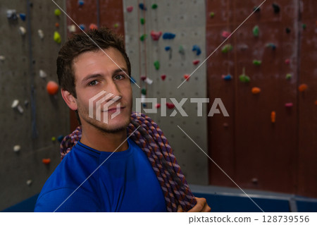 Standing man wearing blue athletic shirt at climbing gym, with rope and colorful holds, copy space 128739556