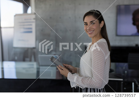 Smiling professional woman holding tablet in modern conference room, with flip chart, copy space Smiling professional woman holding tablet in modern conference room, with flip chart, copy space 128739557