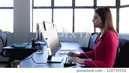 Sitting woman in mid-twenties typing on keyboard at modern office desk, with headset, copy space Sitting woman in mid-twenties typing on keyboard at modern office desk, with headset, copy space 128739558