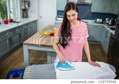 Ironing woman smoothing wrinkles on shirt in modern kitchen, with fruit bowl and iron 128739586