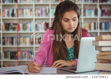 Writing teenage girl studying at library table with open notebook, pen, laptop and stack of books Writing teenage girl studying at library table with open notebook, pen, laptop and stack of books 128739611