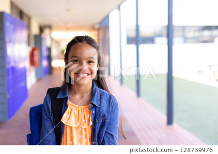 Schoolgirl standing in school corridor, beside blue lockers and fire extinguisher cabinet 128739976