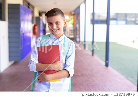 Male child carrying red folder, yellow notebook and blue backpack while smiling in school corridor 128739979