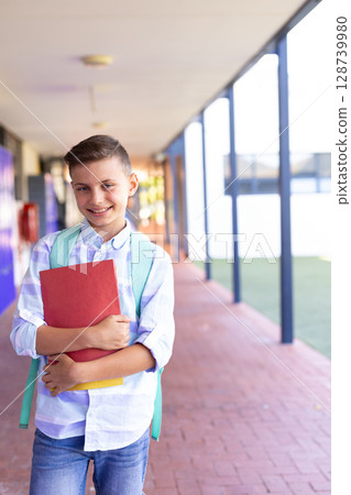 School-age boy walking in school corridor, carrying red binder yellow notebook and green backpack 128739980