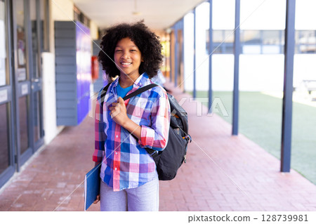 African American boy child standing in school walkway, carrying black backpack, holding blue folder 128739981