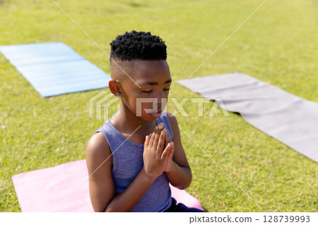 African American boy child kneeling on pink yoga mat in grassy park, meditating with eyes closed African American boy child kneeling on pink yoga mat in grassy park, meditating with eyes closed 128739993