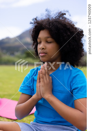 African American boy meditating on pink yoga mat in grassy field with rolling hills 128740009