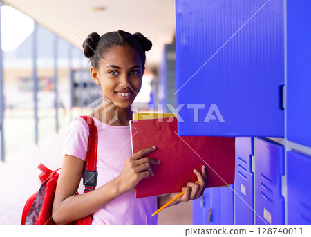 African American teenage girl using locker in school hall carrying red folder notebook red backpack 128740011