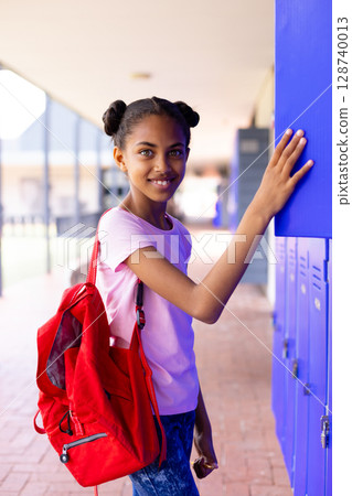 African American girl student smiling at camera in school corridor with red backpack, blue lockers African American girl student smiling at camera in school corridor with red backpack, blue lockers 128740013