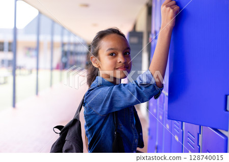 Female child student reaching up to open locker in covered school corridor, carrying black backpack 128740015