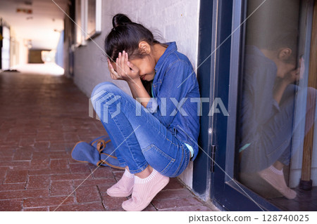 Asian child girl sitting in school corridor hugging knees and crying with blue backpack, copy space 128740025