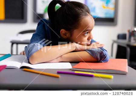 Asian child resting head on desk in classroom, gazing toward maps with notebooks and pencils Asian child resting head on desk in classroom, gazing toward maps with notebooks and pencils 128740028