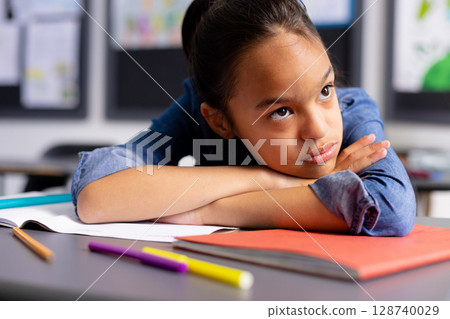 Elementary school girl resting folded arms on desk in classroom, gazing at colored pencils 128740029