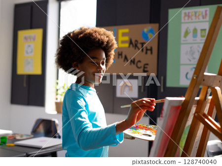 Child boy painting on easel in art classroom with paintbrush, palette and Earth Chance poster Child boy painting on easel in art classroom with paintbrush, palette and Earth Chance poster 128740040