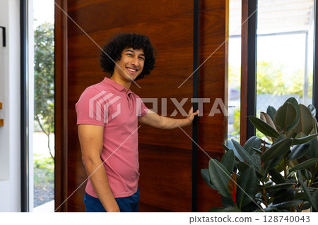 Diverse man opening pivot door in home entryway with rubber plant and glass window showing garden 128740043