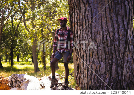 African American man standing on log in forest clearing, wearing red cap and backpack, copy space 128740044