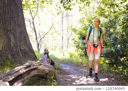 Young woman hiking on trail in sunlit forest with trekking poles, camera, water bottle, copy space 128740045