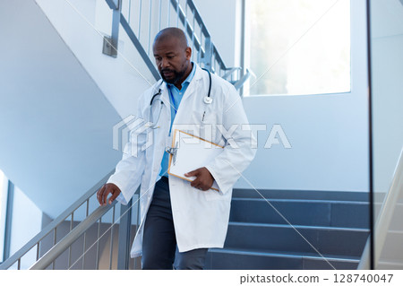 African American male doctor descending hospital stairs, holding clipboard, stethoscope, copy space African American male doctor descending hospital stairs, holding clipboard, stethoscope, copy space 128740047