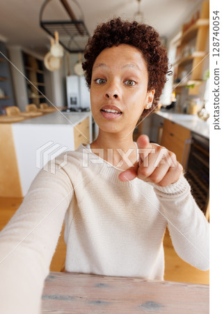 non-binary person in 20s taking selfie and pointing at camera in home kitchen, featuring pot rack 128740054
