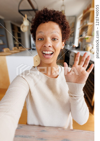 Non-binary person taking selfie in home kitchen on wooden island, holding smartphone 128740055