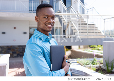 African American man standing in modern courtyard holding laptop under arm, smiling confidently African American man standing in modern courtyard holding laptop under arm, smiling confidently 128740073