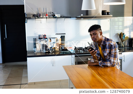 Teenage boy leaning on wooden island counter in kitchen, using smartphone, copy space 128740079