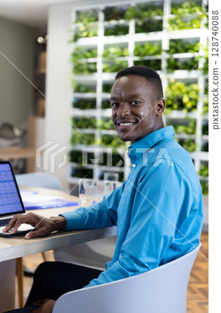 African American man sitting at desk in modern office, using silver laptop with water glasses 128740088