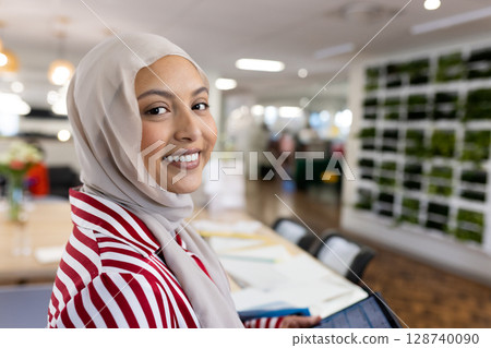 Professional woman wearing hijab working on tablet at coworking table with papers and green wall 128740090