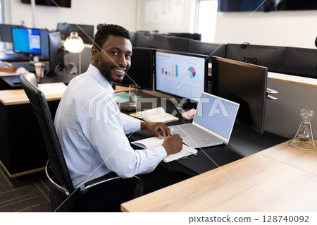 Young African American man writing in notebook smiling at camera at office desk, with laptop 128740092