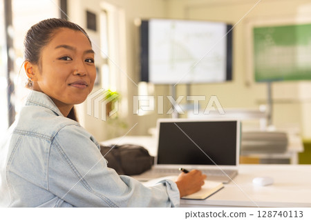 Asian female student writing in notebook at desk in classroom with laptop and screen, copy space Asian female student writing in notebook at desk in classroom with laptop and screen, copy space 128740113