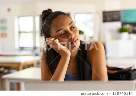 Teenage girl leaning on left hand while gazing at wooden desk in classroom, with open laptop 128740114