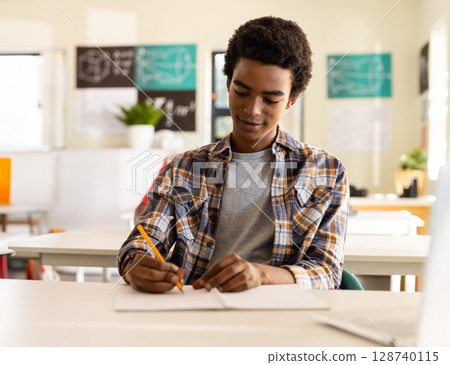 Teenage African American male student sitting in bright classroom, writing notebook with pencil 128740115