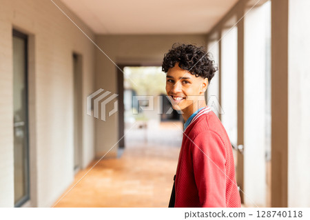 Teenage boy standing and looking back with smile wearing red jacket in campus corridor, copy space Teenage boy standing and looking back with smile wearing red jacket in campus corridor, copy space 128740118
