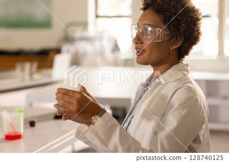 Woman holding clear beaker and explaining experiment in science lab at bench, copy space 128740125